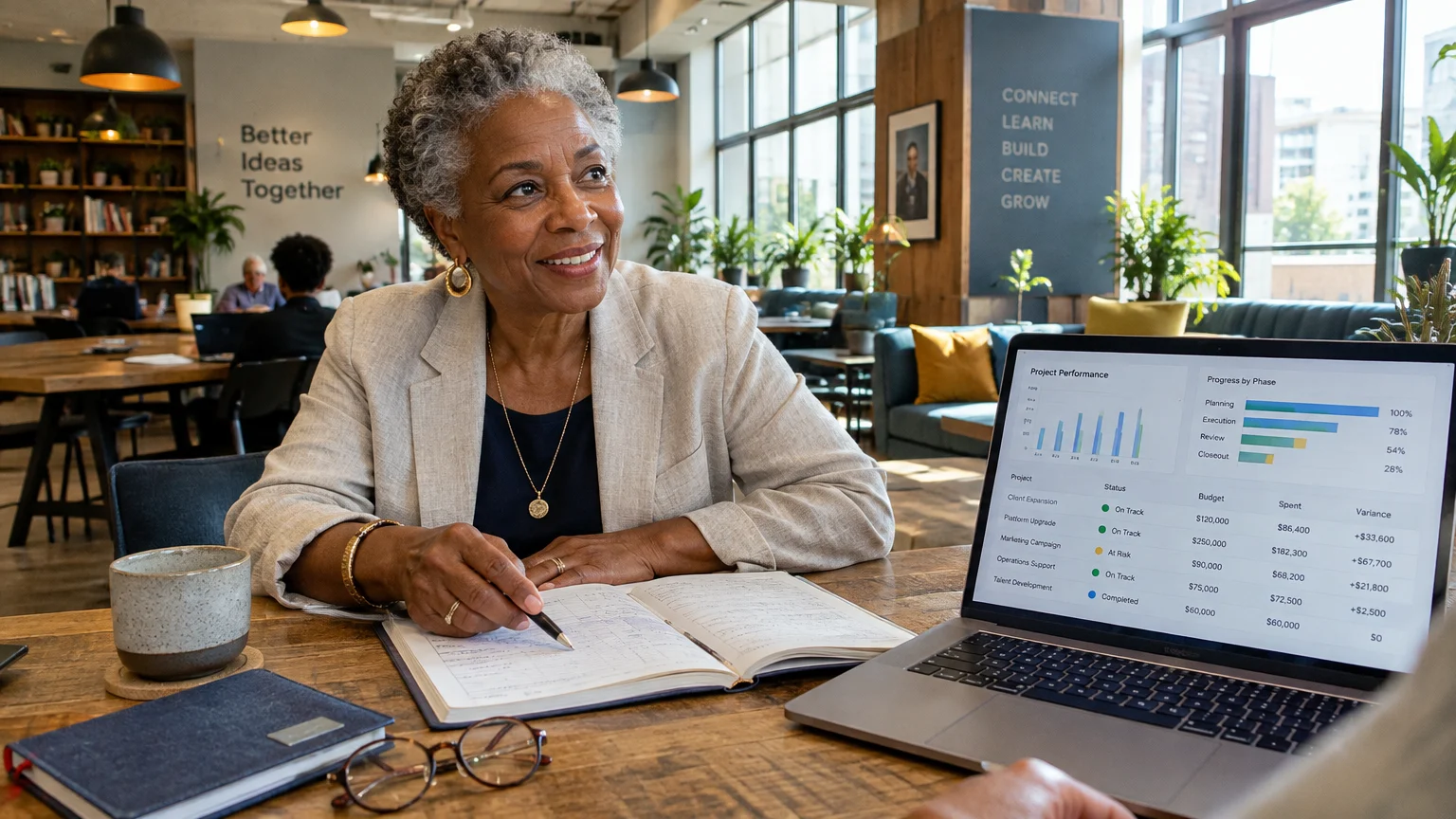 Older woman seated at a desk reviewing a notebook beside an open laptop displaying project management data.